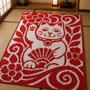 Op de afbeelding: Een rood en wit rechthoekig tapijt met een Japanse Maneki Neko kattenontwerp. De kat zwaait met zijn poot, omringd door bloemen- en krulpatronen. Het tapijt is waarschijnlijk gemaakt van geweven stof.