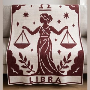 Könnte beinhalten: Eine cremefarbene Decke mit einem bordeauxfarbenen Waage-Tierkreiszeichen-Design. Die Decke zeigt eine Silhouette einer Frau, die Waagen hält, umgeben von Sternen, Blättern und dem Wort "LIBRA".