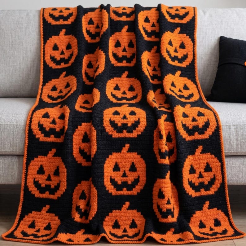May include: A black and orange crocheted throw blanket featuring a repeating pattern of jack-o'-lantern faces. The blanket has a solid orange border and displays multiple orange pumpkin designs with black triangular eyes, noses, and jagged mouths.