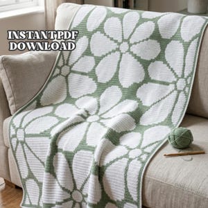 Op de afbeelding: Een wit en groen gehaakte deken met een groot bloemenpatroon ligt over een bank. De deken heeft een madeliefachtig ontwerp. De woorden "INSTANT PDF DOWNLOAD" zijn zichtbaar in de linkerbovenhoek. Een bol groen garen en een haaknaald liggen op de bank.