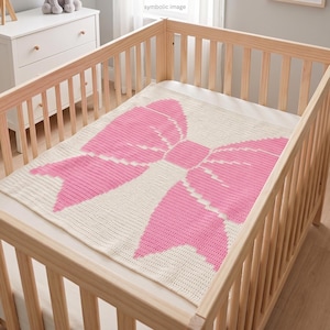 May include: A baby crib with a light wood frame holds a cream-colored blanket. The blanket features a large, pink bow design. The crib is set against a neutral background, suggesting a nursery setting.
