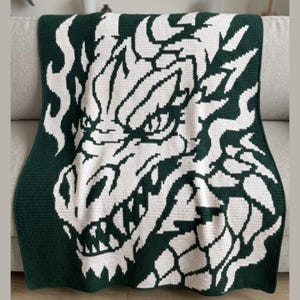 May include: A rectangular crochet blanket featuring a large, detailed graphic of a dragon head in a two-tone color scheme. The design uses dark forest green and off-white yarn to create a high-contrast, pixelated image of the creature's face.