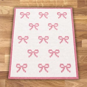 May include: A white rectangular rug with a pink border and a pattern of pink bows. The bows are evenly spaced across the rug's surface. The rug is placed on a wooden floor.