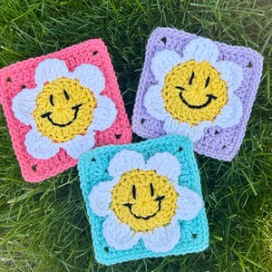 May include: Three crochet squares in pink, light blue, and lavender, each with a daisy design. The daisies have a yellow smiling face centre, white petals, and a coloured square border. The squares are displayed on green grass.