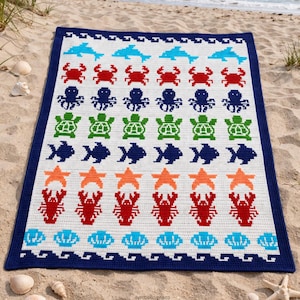 May include: A white crocheted blanket with a navy blue border, featuring rows of sea creatures in various colors. The design includes blue dolphins, red crabs, green turtles, orange starfish, and red lobsters. The blanket is on a sandy beach.