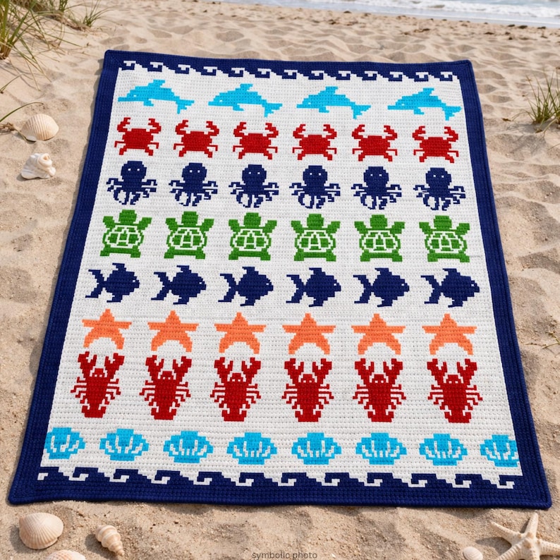 May include: A white crocheted blanket with a navy blue border, featuring rows of sea creatures in various colors. The design includes blue dolphins, red crabs, green turtles, orange starfish, and red lobsters. The blanket is on a sandy beach.