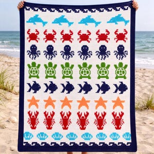 May include: A white and navy blue crocheted blanket featuring a repeating pattern of sea creatures. The design includes blue dolphins, red crabs, green turtles, orange starfish, and red lobsters. The blanket is held up against a beach backdrop.