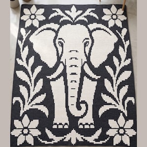 May include: A rectangular rug with a black background and a white elephant design in the centre. The elephant is surrounded by white floral and leaf patterns. The rug is likely made from a woven material, and the design is symmetrical.