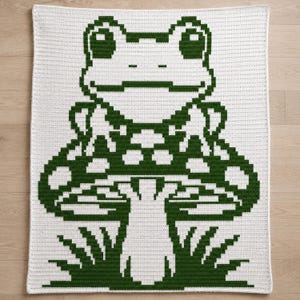 May include: A white crocheted blanket featuring a large, pixelated green frog design. The frog is sitting on a mushroom with grass at the bottom. The blanket is rectangular and has a textured appearance.