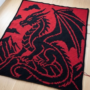 Puede incluir: Manta de ganchillo hecha a mano con un diseño de dragón negro sobre un fondo rojo. El dragón se representa con alas extendidas y un cuerpo detallado. La manta es cuadrada y parece estar hecha de hilo suave.