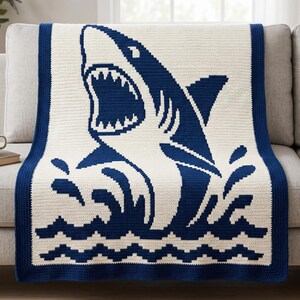 Può includere: Una coperta rettangolare all'uncinetto con una grande grafica di uno squalo blu scuro a bocca aperta su uno sfondo color crema. Il design include onde stilizzate nella parte inferiore e un bordo blu scuro a tinta unita che incornicia il pezzo.