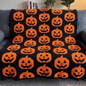 May include: A rectangular crochet blanket featuring a repeating pattern of orange jack-o'-lanterns against a solid black background. The design consists of multiple rows of pixelated pumpkin faces with triangular eyes and jagged mouths.