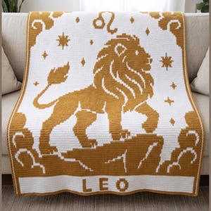 May include: A white and gold crocheted blanket featuring the Leo zodiac sign. The blanket displays a gold lion, stars, and the word "LEO" at the bottom. The blanket is draped over a beige sofa.