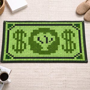 Puede incluir: Una alfombra rectangular verde con un diseño de billete de dólar pixelado. La alfombra presenta dos grandes signos de dólar a cada lado de una imagen central pixelada. El borde es negro y el diseño recuerda a los gráficos de videojuegos retro.
