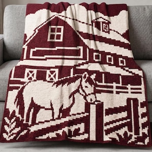 May include: A maroon and cream crochet blanket featuring a pixelated farm scene. The design displays a large barn with a cupola, a fenced-in area, and a horse standing in the foreground. The pattern is created with a repeating grid-like texture.