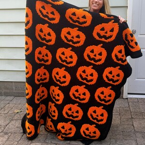 May include: A rectangular crochet blanket featuring a repeating pattern of orange jack-o'-lanterns with black facial features set against a solid black background. The textile displays a grid of pumpkins with triangular eyes and jagged mouths.