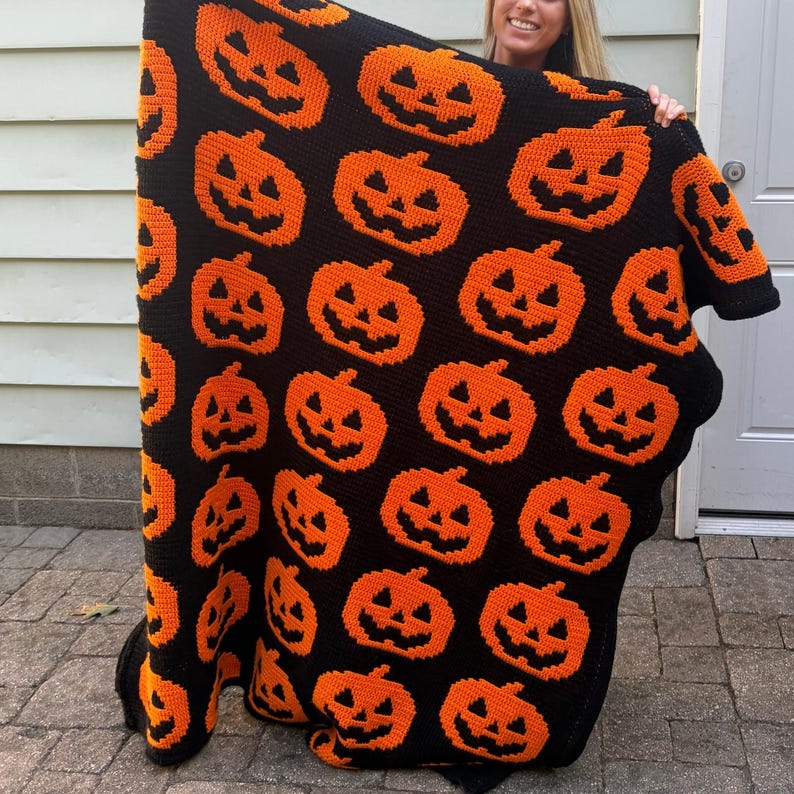 May include: A rectangular crochet blanket featuring a repeating pattern of orange jack-o'-lanterns with black facial features set against a solid black background. The textile displays a grid of pumpkins with triangular eyes and jagged mouths.