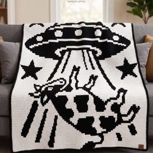May include: A black and white crochet blanket featuring a UFO design with a cow being abducted by a tractor beam. The blanket has a black border and two black star accents. The blanket is draped over a gray sofa.