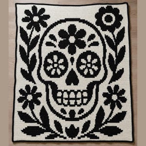 Op de afbeelding: Een gehaakte deken met een zwart-wit suikerschedelontwerp. De schedel is omgeven door bloemen- en bladmotieven. De deken heeft een zwarte rand en is gemaakt van een zacht, getextureerd garen.