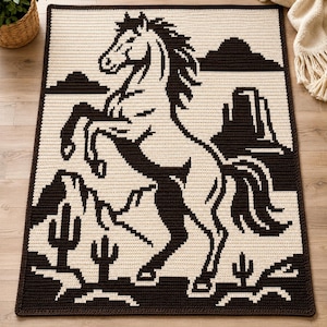 May include: A rectangular crochet blanket featuring a dark brown and cream color scheme. The design depicts a rearing horse in the center, surrounded by a desert landscape with mountains, rock formations, and several small cacti at the bottom.