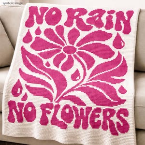 Op de afbeelding: Een witte gehaakte deken met een groot roze bloemmotief en de woorden "NO RAIN NO FLOWERS" in roze. De deken is over een beige bank gevouwen, wat de zachte textuur en de opvallende grafische elementen benadrukt.