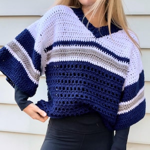 Pode incluir: Um suéter de crochê feito à mão com um design de blocos de cores. O suéter apresenta um decote em V e mangas curtas. O esquema de cores inclui listras brancas, azul marinho e cinza. A parte inferior do suéter tem um design rendado.