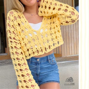 May include: A yellow, open-weave crochet sweater with long sleeves and a V-neck, paired with blue denim shorts. The cropped sweater showcases a handmade design. The image highlights the texture and colour of the garment. The brand logo is visible in the bottom right corner.