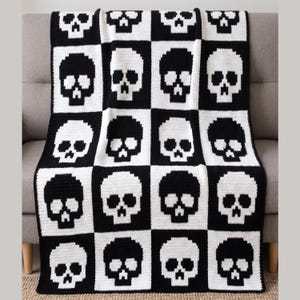 Pode incluir: Um cobertor de crochê preto e branco com um padrão de caveira repetido. As caveiras estão dispostas em um design de tabuleiro de xadrez, com quadrados pretos e brancos alternados. O cobertor está sobre um sofá cinza.