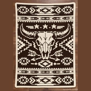 May include: A brown and cream-colored crocheted blanket with a Southwestern design. The blanket features a central bull skull motif, surrounded by geometric patterns and star accents. The border is cream-colored.