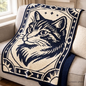 May include: A navy blue and cream-colored knitted throw blanket featuring a detailed cat face design. The blanket is draped over a gray sofa, showcasing a geometric border and a cozy, textured appearance. The words "symbolic photo" are in the bottom right corner.