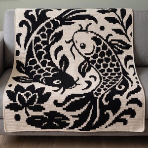 May include: A crocheted blanket featuring a black and cream koi fish design. The blanket displays two koi fish swimming in opposite directions, surrounded by floral and leaf patterns. The blanket is draped over a gray sofa.