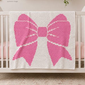 May include: A white and pink crochet baby blanket with a large bow design. The bow is a vibrant pink color, and the blanket is draped over a white crib. The blanket is made of soft yarn and is perfect for a baby.