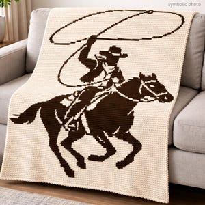 Peut inclure: Couverture en crochet crème avec une silhouette marron d'un cow-boy à cheval, lançant un lasso. La couverture est drapée sur un canapé gris. Les mots "photo symbolique" sont dans le coin supérieur droit.