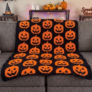 May include: A rectangular crochet blanket featuring a repeating pattern of orange jack-o'-lantern faces against a solid black background. The design consists of multiple rows of stylized pumpkin motifs with triangular eyes and jagged mouths.