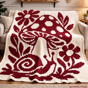 May include: A cream-colored crochet blanket with a large burgundy mushroom, flowers, and snail design. The blanket is draped over a sofa, showcasing the detailed, pixelated pattern. The blanket is a cozy home decor item.