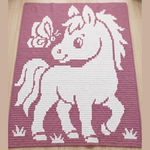 May include: A handmade crochet blanket featuring a white horse and butterfly design on a mauve background. The blanket has a textured appearance and includes grass-like details at the bottom. This item is suitable for a baby or child's room.
