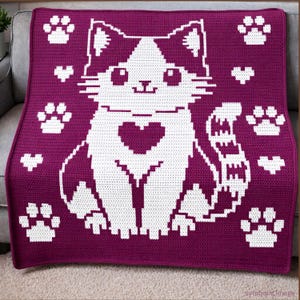May include: A crocheted blanket in a deep purple colour features a large white cat design with a heart on its chest. White paw prints and hearts are scattered across the blanket. The cat has a smiling face.