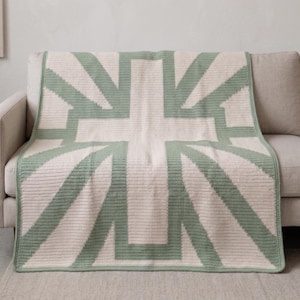 May include: A cream-colored throw blanket with a large, geometric design in light green. The pattern resembles a stylized cross or flag, with thick lines creating the shape. The blanket is draped over a beige sofa, showcasing its texture and design.