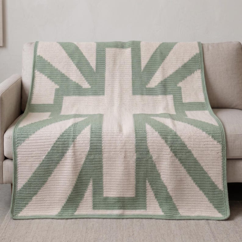 May include: A cream-colored throw blanket with a large, geometric design in light green. The pattern resembles a stylized cross or flag, with thick lines creating the shape. The blanket is draped over a beige sofa, showcasing its texture and design.