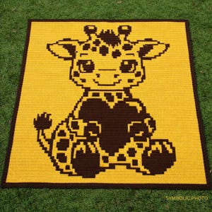 Op de afbeelding: Een handgemaakte gehaakte deken met een glimlachend giraffendesign. De deken heeft een gele achtergrond met een bruine giraffe en een donkerbruine rand. De giraffe houdt een hart vast.