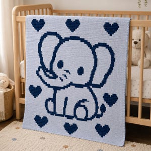 May include: A light blue crochet baby blanket with a navy blue elephant and heart design. The blanket is draped over a wooden cot. A stuffed teddy bear and sheep are in the background.
