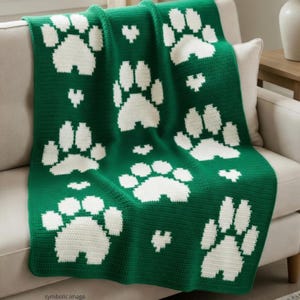Puede incluir: Una manta verde tejida a ganchillo con un patrón de grandes huellas de patas blancas y pequeños corazones blancos. La manta está sobre un sofá beige, mostrando el diseño y la textura del artículo hecho a mano.