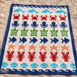 May include: A rectangular beach towel with a white background and a blue border. The towel features a repeating pattern of pixelated sea creatures in various colors, including blue dolphins, red crabs, and green turtles.