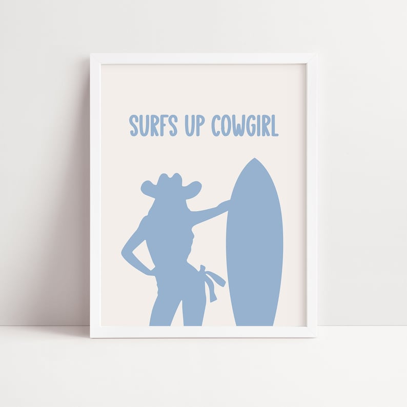 May include: A white framed print with a light blue silhouette of a cowgirl holding a surfboard. The text "SURFS UP COWGIRL" is printed in light blue at the top of the print.