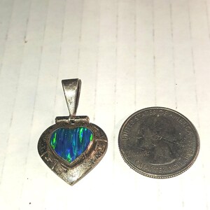 May include: A silver heart-shaped pendant with a vibrant blue and green opal center. The pendant is next to a U.S. quarter for size comparison. The opal has a shimmering, iridescent quality, and the silver setting has an ornate design.