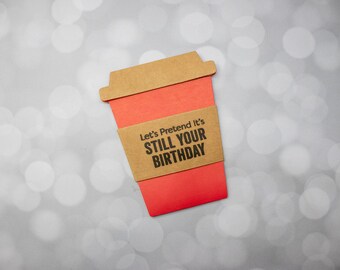 May include: A red paper coffee cup with a brown lid and sleeve. The sleeve reads "Let's Pretend It's STILL YOUR BIRTHDAY" in black text. The background is a soft, out-of-focus, bokeh effect.
