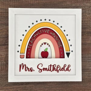 May include: A white framed wooden sign featuring a rainbow design with the words "Teach Love Inspire". The rainbow is made of layered wood in shades of red, pink, and yellow. A red apple is in the center. The name "Mrs. Smithfield" is below the rainbow.