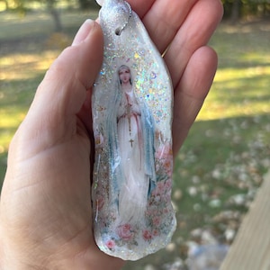 Puede incluir: Un colgante religioso con una imagen de la Virgen Mar&iacute;a, adornado con purpurina y un cord&oacute;n plateado. El colgante tiene una base blanca con una imagen impresa de Mar&iacute;a con una t&uacute;nica blanca y velo azul, rodeada de rosas rosas.