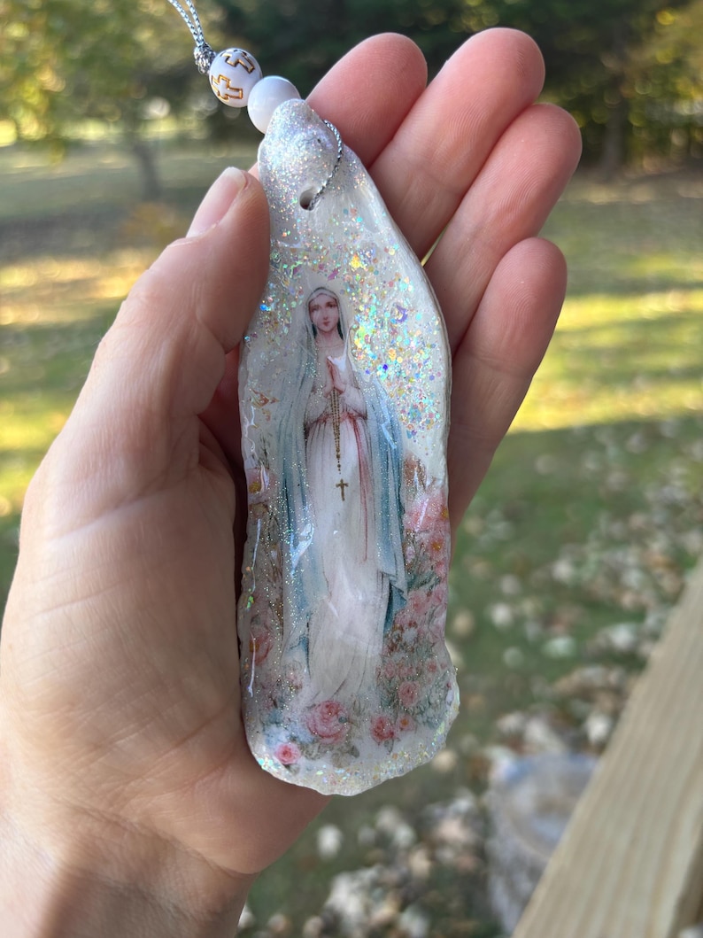 Puede incluir: Un colgante religioso con una imagen de la Virgen Mar&iacute;a, adornado con purpurina y un cord&oacute;n plateado. El colgante tiene una base blanca con una imagen impresa de Mar&iacute;a con una t&uacute;nica blanca y velo azul, rodeada de rosas rosas.