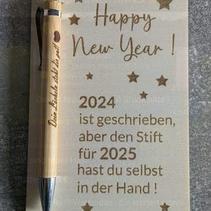 May include: A wooden sign with the words "Happy New Year!" and "2024" engraved, accompanied by star designs. A bamboo pen with a silver tip and the words "Dein L&auml;cheln steht dir gut!" is placed on the sign.